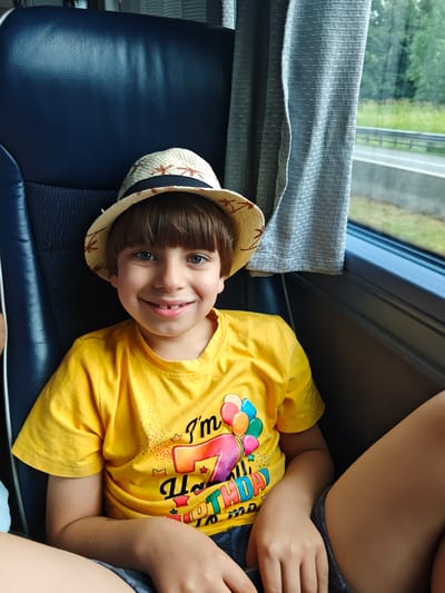 The image shows a young boy wearing a straw hat and a yellow t-shirt with a "Happy Birthday" graphic. He is sitting on a bus or train, with a blue seat behind him and a window to his right. The window shows a view of trees and a road, suggesting a daytime setting. The boy is smiling broadly, revealing a gap where a tooth is missing. The t-shirt has balloons and the number "7" printed on it, along with the words "I'm 7" and "Happy Birthday." The overall mood of the image is cheerful and celebratory. The location is identified as Eugendorf, Austria.