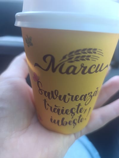 The image shows a close-up of a person's hand holding a yellow paper coffee cup. The cup has a white lid and features brown text and graphics. The text reads "Marcu Savurează trăiește, iubește!". Above the text, there's a stylized graphic of wheat stalks. There's also a small, green butterfly graphic on the left side of the cup and a pink flower graphic below the butterfly. The background is blurred, suggesting the photo was taken outdoors or in a moving vehicle. The lighting is bright, indicating daytime. The overall impression is casual and perhaps related to enjoying a drink. The location context is Pohorniceni, Moldova.