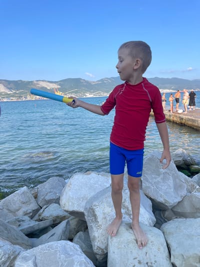 The image depicts a young boy standing on large rocks next to the sea, likely in Ol'ginskii, Russia. He is wearing a red rash guard and blue swim trunks, and holding a blue and yellow toy, possibly a water gun or floatation device. The boy is looking down towards the water.

In the background, several people are visible standing on a concrete walkway by the sea. Beyond the walkway, there is a town or city nestled against a backdrop of mountains. The sky is a clear blue, suggesting fair weather and likely daytime. The sea is calm with ripples on the surface. The overall scene conveys a sense of a vacation or day at the beach.

There is visible text on the boy's swim trunks: "ZOGGS".