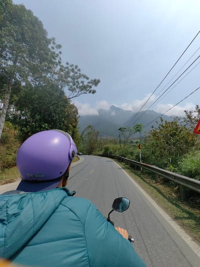 The image presents a first-person perspective from a motorcycle traveling on an asphalt road in the city of Bình Lư, Vietnam. The back of a person, identified as the rider or passenger, occupies the foreground, wearing a purple full-face helmet and a teal quilted jacket. Their right hand is visible on the motorcycle's handlebar, with a rearview mirror partially reflected. The two-lane road features a solid yellow center line and a solid white fog line along its right edge, curving gently to the left in the distance. A metal guardrail parallels the right side of the road. Immediately beyond the guardrail, a yellow and black chevron road sign indicates a left curve, positioned near a partially visible red and yellow triangular warning sign. Power lines are visible stretching across the upper right sky. The surrounding environment consists of dense green vegetation and trees lining both sides of the road. In the background, multiple mountain peaks, partially obscured by clouds or atmospheric haze, rise under a clear to slightly hazy blue sky. A white car is visible on the road ahead, approaching the curve.