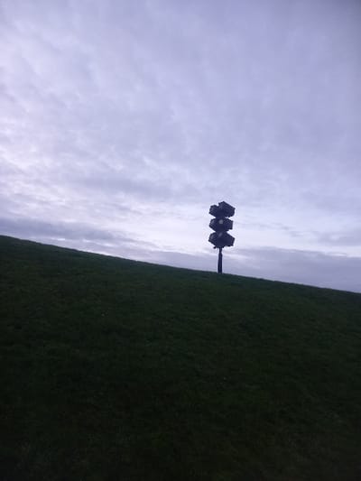 A vertical, eye-level shot captures a dark object, potentially a street light or antenna, silhouetted against a cloudy sky and verdant grass. The clouds fill the majority of the frame, their varying shades of grey contrasting with the solid green of the grass, which stretches across the right side of the image. The dark object pierces the horizon line, a stark figure against the muted backdrop.