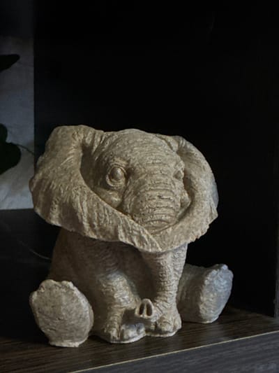 A tan stone elephant figurine sits on a dark surface with a dark background. The figurine has textured skin and is facing towards the right. The surface it is on has a dark wood-grain edge. The location in Kladovitsy, Russia, is not discernible from the image.