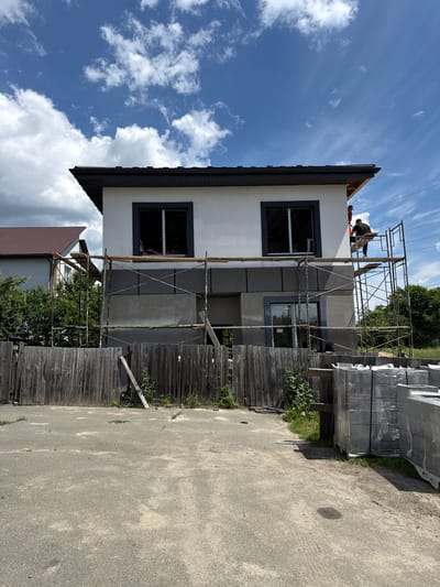 The image depicts an exterior view of a multi-story building under construction in Bucha, Ukraine. Two individuals are visible on the roof of the building, engaged in work. Scaffolding surrounds a section of the building's facade, indicating ongoing construction or renovation activities.
The scene is set outdoors under a bright, partly cloudy sky, suggesting daytime. A pile of materials, likely construction supplies, is stacked to the right of the building, wrapped in plastic. A wooden fence runs along the side of the property, with some overgrown vegetation visible. The building itself has a light-colored exterior with dark-framed windows.
The overall impression is one of active construction work in a residential or semi-rural setting. There is no visible text in the image.