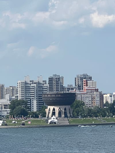 The image captures a waterfront view of a city skyline, likely on a sunny day with scattered clouds. In the foreground, a body of water, possibly a river or lake, is visible with ripples on its surface and two small boats moving across it.
The midground features a grassy embankment with trees and a paved walkway lined with lights. Several tents or temporary structures are set up on the lawn, suggesting an event or gathering. A distinctive building with a large, dark, bowl-shaped roof supported by arched structures stands prominently in the center of the scene. Behind this unique structure, a cluster of modern, multi-story buildings forms the city's skyline. These buildings are predominantly white and gray, with visible windows and balconies.
The background consists of a pale blue sky with white, fluffy clouds. The overall scene suggests a lively urban waterfront, potentially a park or recreational area within a city. The specific location of Дачное, Russia is provided as context for this urban landscape.