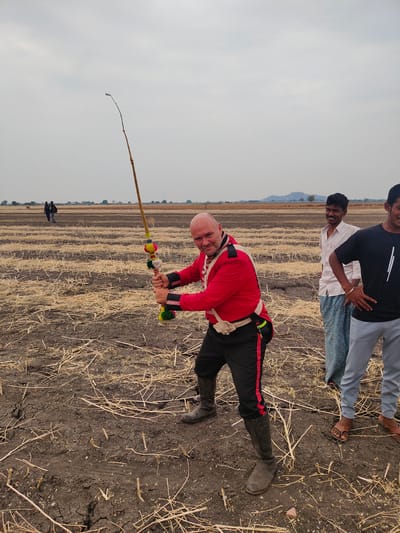 The scene is an open agricultural field in Kottalapalli, India, under an overcast, light grey sky. The ground consists of dry, dark brown soil exhibiting cracks, with scattered light brown crop stubble from a previous harvest.
In the foreground, a bald Caucasian male, appearing to be in his 40s to 50s, stands centrally, facing the camera with a wide smile. He is dressed in a red, uniform-style jacket with black collar and cuff trim, black trousers featuring a red stripe down the side, and dark, knee-high boots. A light-colored pouch is attached to a belt around his waist. He holds a long, flexible, whip-like object with both hands, positioned across his body. This object is primarily striped yellow and brown and is decorated with multiple colorful pom-poms (red, yellow, green, blue) along its length, particularly near the handle.
To the right of the Caucasian male, two Indian males are standing. The first, positioned slightly behind, is a young adult wearing a light-colored, possibly striped, button-up shirt and a blue-and-white patterned lungi. He is smiling towards the man in red. The second, partially visible on the far right, is also a young adult, wearing a black t-shirt and light grey trousers, with his hands on his hips. He is smiling and wears brown sandals.
In the distant background, two indistinct figures are walking across the field on the left. Further to the right in the mid-ground, a group of livestock is visible. The horizon features low hills and a few scattered trees.