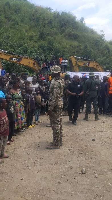 A group of people stand in front of two yellow Caterpillar excavators. The excavators are parked on a dirt road in front of a hill covered in green vegetation. The group is composed of men, women, and children. The adults are wearing a variety of clothing, including uniforms, casual clothing, and traditional clothing. The children are wearing a variety of clothing, mostly casual. The people are gathered around a white banner with blue lettering. The scene is set in Sake, Democratic Republic of Congo, at coordinates -1.571, 29.046. A man wearing a camouflage uniform, and a black hat with a camouflage strap is facing the viewer. He is in the center of the group. A man in a black suit and a man in a camouflage uniform with a black cap are standing to the right of the man in camouflage. A group of children are standing in front of the men in black and camouflage uniforms. The children are looking up at the men. The scene is quiet and still. There is no apparent action or interaction between the people. The environment is dusty and dry. The ground is covered in dirt and small rocks. The sky is cloudy. The scene is typical of a rural area in the Democratic Republic of Congo.