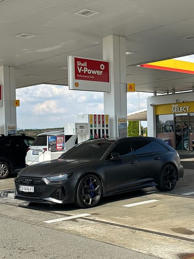 The image shows a matte black Audi RS 6 parked at a Shell gas station in Drazhevo, Bulgaria. The car is in the foreground, facing the viewer. To the left of the Audi, a white SUV with license plate CB 0606 KO is also visible. In the background, there are gas pumps and a Shell Select convenience store. The sky is cloudy.
