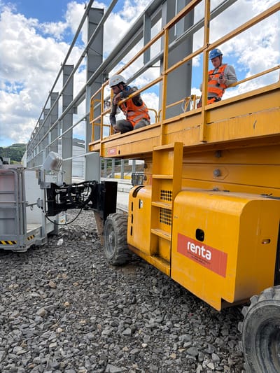 Two construction workers are on a yellow scissor lift, with one worker kneeling and looking down, while the other stands and operates a power tool. The scissor lift is positioned on a surface covered in gravel, adjacent to a large metal construction structure. In the background, there are hills and a cloudy sky. The logo "renta" is visible on the side of the scissor lift, indicating it is a rental company. The scene is set in Janowice, Poland.