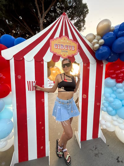 The image depicts a young woman posing in front of a circus-themed booth. The booth has red and white stripes and a pointed roof, adorned with a sign that reads "WELCOME TO THE CIRCUS SHOW." She is wearing a black crop top, a denim skirt, sunglasses, and sandals. In her left hand, she holds a red and white striped popcorn box. The scene is decorated with a large arrangement of blue, white, and red balloons, as well as golden and silver balloons. Bubbles are visible floating in the air, creating a festive atmosphere. The background shows trees and a street, suggesting an outdoor setting. The overall impression is that of a lively event or celebration.