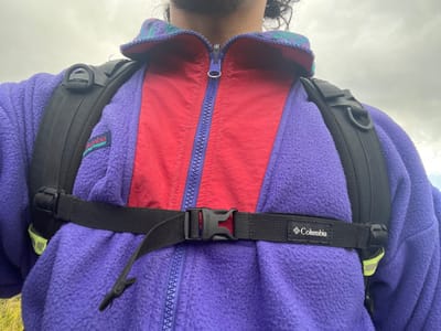 The image displays the upper torso of an individual, seen from a first-person perspective, outdoors at Estancia Harberton, Argentina, under an overcast sky. The subject wears a purple fleece jacket with a patterned collar (teal, green, purple) over a red jacket that features a purple zipper and collar. A "Columbia" brand patch is visible on the left side of the purple fleece. A black backpack is worn, with visible padded shoulder straps, each fitted with a black D-ring. A black webbing sternum strap, fastened by a black plastic buckle, crosses the chest. This sternum strap also features a "Columbia" brand tag on the right side. A fluorescent yellow-green reflective element is partially visible on the right side of the subject's body, possibly on the backpack or an additional layer.