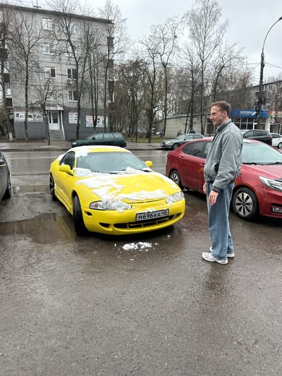 A man stands on wet pavement next to a yellow car covered with snow on its hood and windshield. The car has a license plate number M696KA 46. Another red car is parked behind it. The man is wearing a light grey jacket, jeans, and white sneakers. Bare trees line the street, and a grey building with several signs is visible in the background. The location is the city of Kursk, Russia.