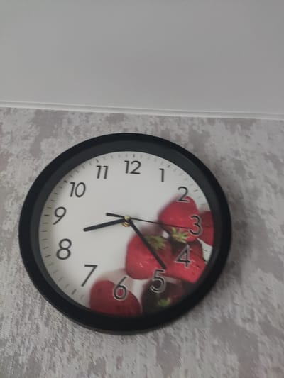 A round analog clock with a black frame is mounted on a textured, light-colored wall. The clock face is white with black numerals and markings. The numbers 1 through 12 are visible, with the exception of the number 12 which is partially obscured. The clock features two black hands, an hour hand and a minute hand, both pointing to the left of the clock face. The clock face is decorated with a pattern of red strawberries. The image does not provide any information or visual cues that would allow for its location to be identified within the city of Gorno-Altaysk, Russia.