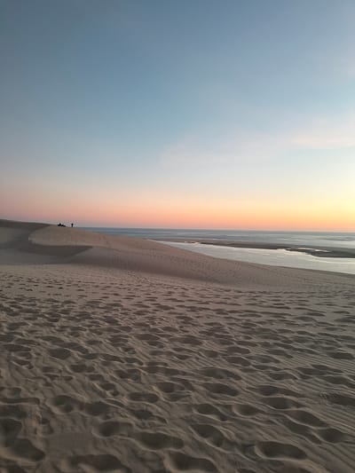 This is a landscape photograph of the Dune of Pilat in La Teste-de-Buch, France, during sunset. The foreground is dominated by rippled sand dunes covered in numerous footprints. In the midground, a line of people can be seen standing and sitting on the crest of a dune, silhouetted against the sky. Beyond the dunes, a body of water, likely the Atlantic Ocean, stretches to the horizon, where the sky transitions from a pale blue at the top to soft hues of pink and orange near the horizon, indicating the setting sun. The overall impression is one of a serene, natural environment at dusk.