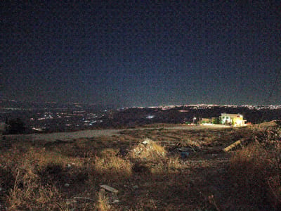 The image depicts a nighttime landscape with a vast, dark sky speckled with noise. Below the sky, a sprawling city lights up a distant horizon, with clusters of illuminated buildings and streets suggesting a populated area. In the middle ground, a solitary, brightly lit building, possibly a house or small establishment, stands out against the darker terrain. The foreground is a rough, uneven landscape covered in dry, sparse vegetation and debris, suggesting an uncultivated or neglected area. The overall impression is one of a semi-rural or elevated viewpoint overlooking a city at night. The provided context indicates the location is in Umm al-Quttayn, Jordan.