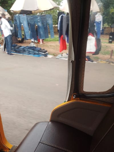 The photo is taken from the back seat of a yellow auto rickshaw. The rickshaw is parked on the side of a paved road with a roadside market visible in the background. The market is selling blue jeans. A person wearing a white shirt and jeans is standing near the jeans in the market. The image is taken in Abuja, Federal Capital Territory, Nigeria at coordinates (latitude: 8.955, longitude: 7.552).