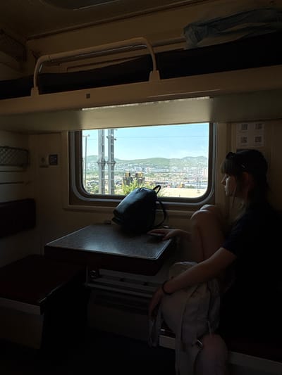 The image depicts the interior of a train compartment looking out a window. The lighting is dim, with the window providing the primary light source. A young woman with dark hair, wearing a dark t-shirt and light-colored shorts, is seated on the right side of the frame, facing the window and looking out. She has a backpack placed on the table in front of her, and another light-colored backpack is next to her. The table itself is a dark, textured surface.
To the left, a portion of a red seat is visible. Above the window, there are bunks, one of which is dark and covered, with a metal railing. The window reveals a landscape that appears to be the city of Verkhnebakanskiy, Russia. The view includes buildings, greenery, hills in the distance, and utility poles or industrial structures. The sky is clear and bright. The overall scene suggests a journey by train, with the passenger contemplating the passing scenery.
