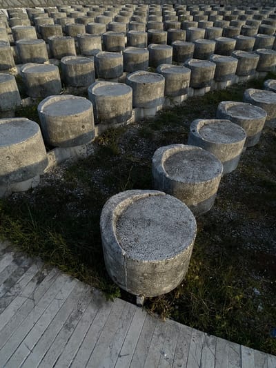 The image depicts an outdoor installation or structure located in Barutana, Montenegro. The scene is dominated by hundreds of grey, weathered concrete cylindrical blocks, uniformly arranged in stepped, linear rows that ascend towards the background. Each cylinder measures approximately 40-50 cm in height and 50-60 cm in diameter. Many of these cylinders feature a distinct C-shaped or crescent-shaped indentation cut into their top surface, creating a partial raised edge around a central flatter area. The concrete surfaces show signs of weathering, including minor chipping and some white speckled discoloration on the top surfaces. The cylinders are primarily supported by smaller, lighter-colored concrete bases, elevating them slightly above the ground level.

The ground between and beneath the concrete blocks consists of sparse, overgrown green grass, weeds, and dark gravelly soil. In the immediate foreground, to the left, a section of light-colored, rectangular paving stones or concrete slabs is visible, forming a horizontal pathway or edge adjacent to the grassy area. Natural daylight illuminates the scene, casting distinct shadows to the right of the concrete cylinders, suggesting sunlight originating from the left. There are no individuals, animals, or active subjects present in the frame.
