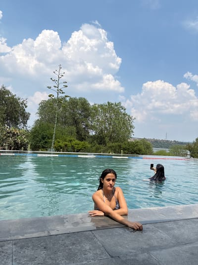 Two women are in an outdoor swimming pool in Pueblo Nuevo de Jasso, Mexico, in daylight. One woman is leaning on the pool's edge, facing the camera. The other woman is in the water, holding a smartphone. The background includes trees and a partly cloudy sky.