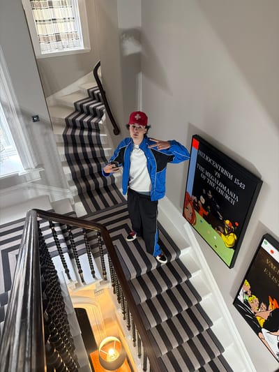 The image captures a person standing on a staircase adorned with a striped runner. The person, wearing a blue and black jacket, black pants, a red cap, and sneakers, is holding a phone in their left hand and posing with their right hand. The staircase features a dark wooden handrail and balusters. Two framed artworks are visible on the wall beside the staircase. The environment appears to be an indoor residential setting, likely within a house. The location is identified as Culross, United Kingdom, though no specific landmarks within the city are depicted.