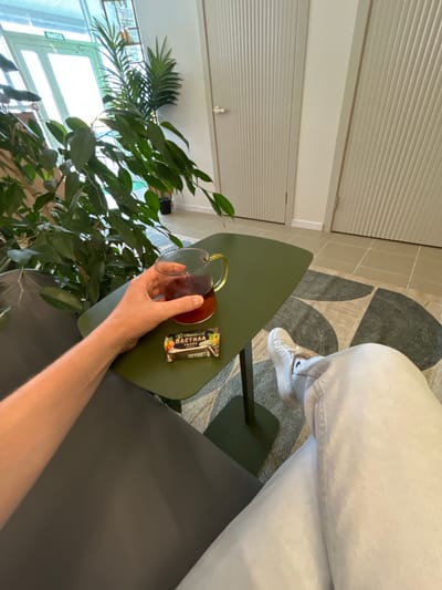 A first-person perspective shot captures a person's arm holding a glass of dark liquid on a green side table. Next to the glass, a small candy bar with an illustration of fruit is visible. The background reveals an indoor environment with large potted plants, two cream-colored doors with vertical ribbed detailing, and a rug with a circular pattern on the floor. The scene is illuminated by natural light coming from a glass-paneled exit sign.