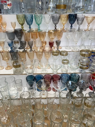 The image shows a large collection of glassware displayed on a white surface, possibly a market stall in Eshanguzar, Uzbekistan. The glassware includes various types of glasses, goblets, and bottles, many with decorative patterns and colored tints. Some glasses are clear, while others are in shades of blue, green, amber, and ruby.
The items are arranged in rows, with some upright and others tilted. The lighting suggests it is daytime. The scene is an arrangement of merchandise, likely for sale. There are no people visible in the image.
On one of the bottles on the far left, a label is visible with text that appears to be in Russian and possibly English, with the word "WINXX" and what looks like "ORANGE" or a similar word. The overall impression is of a vibrant display of glassware.