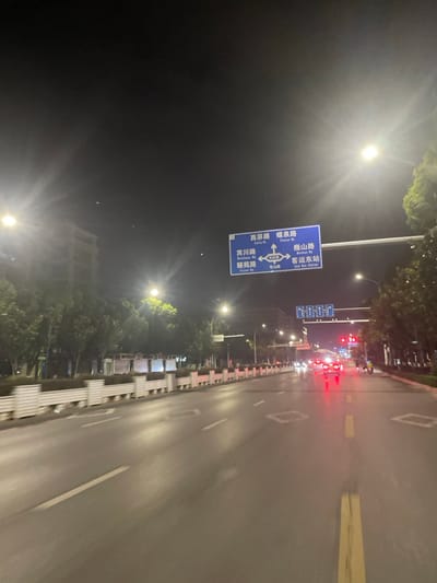 This night-time image depicts an urban street scene in Dali City, Yunnan Province, China, specifically at or approaching an intersection involving Binchuan Road, Weishan Road, and routes leading to the Guest Bus Station, as indicated by overhead signage. The scene is illuminated by numerous bright streetlights and vehicle lights.

The foreground features a wide, multi-lane asphalt road, appearing somewhat reflective, potentially from light rain or a wet surface, marked with solid white lane lines, dashed white lines, a double yellow center line, and square-shaped road markings. To the left, a median or pedestrian barrier comprised of white, rectangular planters filled with green shrubbery separates the road from a tree-lined area. Further left, the indistinct outlines of multi-story buildings are visible.

Centrally positioned over the road is a large, blue overhead gantry sign displaying white text and arrows. The sign indicates directions for "Xiping Rd (西屏路)", "Dieqian Rd (蝶泉路)", "Binchuan Rd (宾川路)", "Weishan Rd (巍山路)", "Lijiang Rd (丽江路)", "Tianbo Rd (天波路)", and "Guest Bus Station (客运站)".

Further down the road, a set of overhead traffic lights is visible, displaying a red signal. Approximately three to four vehicles are present in the distance, their red taillights brightly illuminated, indicating they are stopped or slowing for the traffic signal. On the far right lane, a single scooter or motorcycle is also visible, displaying a red taillight. No individuals are clearly discernible on foot or as drivers/passengers within the vehicles. The sky above is dark, indicating night.