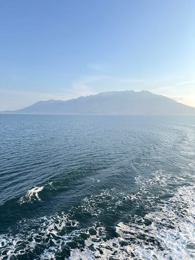 The image displays an expansive maritime and mountainous landscape observed from a moving vessel near Palaiopoli, Greece. The foreground and midground consist of deep blue-green sea water, characterized by numerous surface ripples. A prominent white foam wake extends across the bottom right, indicating the motion of the viewing platform. A distinct, white-crested wave is visible in the lower-left quadrant. Horizontally spanning the background is a large mountain range, its upper elevations exhibiting patches of snow. The mountains appear somewhat diffused by atmospheric haze. The sky above is clear and light blue, with minimal cloud cover or atmospheric features, suggesting daylight conditions. No human subjects or specific artificial structures are visible, aside from the implied vessel creating the wake.