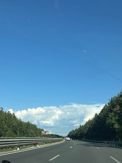 This is a vertical, wide-angle photograph taken from a vehicle traveling on a highway. The scene is dominated by a bright blue sky with scattered white clouds. To the right of the frame, a dense line of green trees borders the highway, creating a natural barrier. The highway itself is asphalt with white lane markings and a metal guardrail. Several vehicles are visible on the road ahead, including a white car and a red car. The time of day appears to be daytime, with clear and sunny weather. The overall impression is one of a scenic drive through a forested area. A billboard is visible on the left side of the highway, featuring text and an image of a person. The location is identified as Novi Han, Bulgaria.