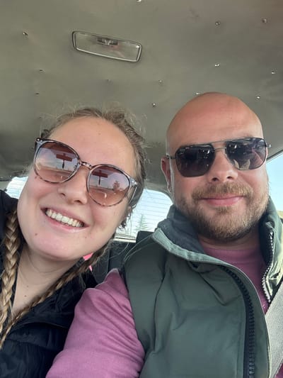 The image displays a selfie taken from within a vehicle, featuring two individuals, a female and a male, in what is identified as the city of Chintulovo, Bulgaria. The female subject, positioned on the left, has light-colored hair, part of which is braided, and is smiling with visible teeth. She wears large, round, tortoiseshell-patterned sunglasses and a dark-colored top. The male subject, positioned on the right, is bald with a light-brown beard and mustache, and is smiling subtly. He wears aviator-style sunglasses, a long-sleeved pink or maroon shirt, and a dark olive-green puffer vest with a visible black zipper. A seatbelt strap crosses his right shoulder.
The interior background shows the vehicle's light-colored headliner with several small indentations or fasteners, and a rectangular rearview mirror is mounted centrally above the subjects. Through the rear window, a bright outdoor sky and the tops of green trees are visible. Reflections in both subjects' sunglasses display an outdoor scene with trees and what appears to be a vertical pole or sign structure. Both individuals are looking directly towards the camera, suggesting a posed photograph.