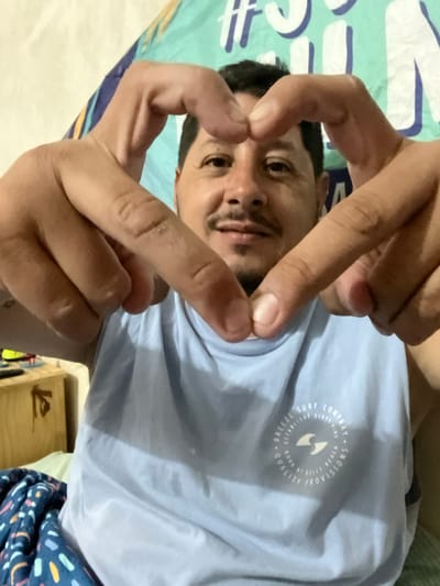 An adult male, approximately 30-45 years old, with dark hair, a mustache, and a light goatee, is depicted in a medium close-up, smiling faintly and looking directly at the camera. He is forming a heart shape with both hands, positioned in front of his face. He wears a light blue sleeveless shirt featuring a white circular logo on the chest that reads "BREEZE SURF COMPANY ESTABLISHED SIXTY-EIGHT BORN AT KEELYS BEACH QUALITY PROVISIONS" around a stylized 'S' or wave emblem. The background consists of a patterned turquoise and white fabric or tapestry behind his head. To the lower left, a dark blue patterned blanket is visible, and further left, a light-colored wooden nightstand with indistinct objects on top can be seen. The scene is an indoor setting in Bernal Este, Argentina.