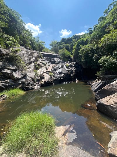 A wide, eye-level shot captures a natural scene at Hoa Thang Ward, Vietnam, dominated by a rocky waterfall and a pool of water. The sky above is a vibrant blue, dotted with white, fluffy clouds. Lush green vegetation covers the steep, rocky slopes on either side of the waterfall.

The waterfall itself is composed of dark, jagged rocks, with water flowing down in a thin stream visible in the upper right portion of the frame, disappearing into the shadows of a small cave-like opening. At the base of the waterfall, a dark, calm pool of water reflects the surrounding foliage and sky. The water is murky green, with visible sediment and debris near the edges.

In the foreground, to the left of the pool, a patch of bright green grass grows on a rocky outcrop. The rocks in the foreground are large and varied in color, ranging from dark gray to reddish-brown. Some of these rocks are partially submerged in the shallow edges of the pool. The overall impression is one of a secluded, natural environment. No people or animals are visible in the scene.