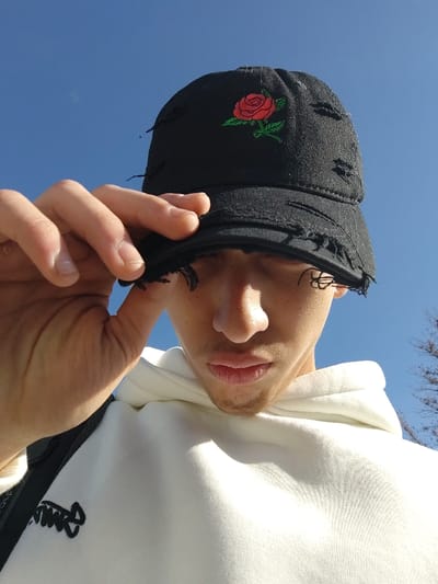 A young adult male is depicted in a close-up, low-angle shot against a clear blue sky. He wears a black, distressed baseball cap with an embroidered red rose, green stem, and leaves on the front panel. His right hand is raised, with fingers gripping the distressed brim of the cap, partially obscuring his upper face and eyes. A white hooded sweatshirt with a black embroidered design or text on the left chest area is visible on his torso. The lower portion of his face, including his nose, mouth, and chin with light facial hair, is visible. In the lower right background, dark, bare tree branches are faintly visible against the sky. The lighting suggests bright daylight conditions.