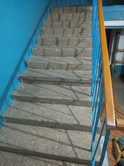 The image shows an indoor concrete staircase with a bright blue wall on the left and a blue metal railing with a wooden handrail on the right. The stairs are lit by artificial light, casting shadows from the railing onto the steps. The concrete of the steps is speckled with lighter-colored aggregate. In the background, a portion of a doorway and some construction materials are visible. The scene appears to be in a utilitarian building, possibly a stairwell in an apartment complex or an industrial setting. There is no visible text in the image. The time of day and weather are not determinable from the image. The overall impression is one of an everyday, functional space.