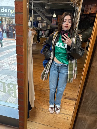 A female individual is depicted in a full-length mirror selfie, positioned inside a retail establishment in Belfast, United Kingdom. She stands on a polished wooden floor, facing the mirror directly. The individual has dark hair, and white horizontal strips of face paint are visible beneath her eyes and on her upper cheeks. She is wearing a dark leather jacket over a short-sleeved green jersey shirt with the white text "TEAM" visible across the chest. Her midriff is exposed above light blue jeans featuring a distinctive V-shaped open waistline. A green and pink plaid scarf is draped around her neck. On her feet are white and brown patterned clogs with sheepskin lining. She holds a smartphone with a pink case and a triple camera system in her right hand, angled towards the mirror to capture the reflection. A black camera body is slung over her left shoulder, and a blue bag is also visible over the same shoulder.
The retail interior reflected in the mirror includes multiple clothing racks displaying garments, a staircase with dark railings in the background, various posters on the walls, and industrial-style pendant light fixtures.
To the left of the mirror, an exterior urban scene is reflected. This includes a tiled sidewalk and the facade of an adjacent building featuring a sign partially readable as "Goldenpl..." and "Belfast." A male individual wearing a blue puffer jacket and green trousers is seen walking on the sidewalk in the reflection.