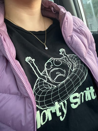 A person is wearing a partially zipped lavender puffer jacket and a black T-shirt with a white cartoon graphic and the words "Dirty Smit" printed on it. A gold chain necklace with a pendant is visible around the person's neck. The scene appears to be inside a vehicle, possibly a car, due to the presence of a window and part of a car interior visible in the background. The location in the city of Santiago de Surco, Peru, is not discernible from the image.