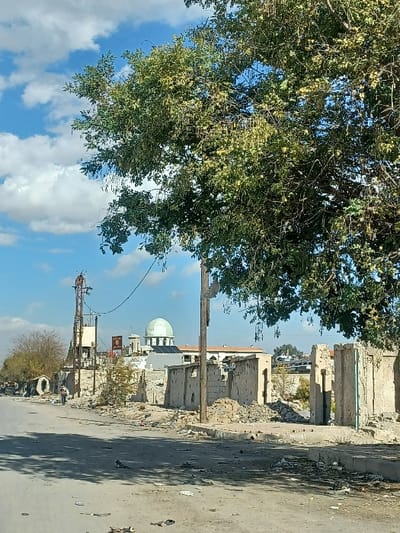 The image displays a daylight street scene in Al-Qadam Municipality, Syria, under a partly cloudy blue sky. A large, leafy tree with light green foliage and visible seed pods occupies the upper right and central-right portions of the frame, partially obscuring the sky and distant elements.

In the foreground, a paved road surface shows significant wear, dirt, and scattered debris including plastic and paper. Shadows are cast across the road from unseen objects to the left.

The mid-ground reveals extensive urban destruction. On the right side of the road, multiple structures appear heavily damaged or partially demolished, consisting primarily of exposed concrete or stone walls with visible impact holes and areas of missing material. Piles of rubble lie at their bases. A wooden utility pole stands prominently among these damaged structures.

On the left side of the street, further back, another damaged building is visible. A tall, metallic utility pole with cross arms and electrical wiring stands near the left edge of the frame. An individual, wearing dark clothing, is walking away from the viewer on the left side of the road in the distance, partially obscured by the environment.

Centered in the mid-ground, a prominent mosque-like structure is visible. It features a large, light green dome atop a white building, flanked by smaller domes or architectural features. To the left of the mosque, a red and black sign or billboard with a portrait is mounted. Additional damaged buildings and general debris fill the area between the foreground and the mosque. Vehicles or large objects are faintly discernible in the far background.