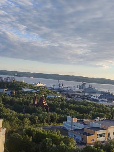 This is a high-angle, vertical, full shot of a coastal city. The sky is filled with scattered clouds. In the middle ground, a bay is visible, with several large ships docked along the shore. The city itself appears to be built on rolling hills covered in lush green trees. A prominent, reddish-brown church with golden domes stands out against the greenery. In the foreground, modern buildings with grey and tan facades are visible. The overall scene suggests a harbor town with a blend of historical and contemporary architecture, situated in a natural, hilly environment. The location is Severomorsk, Russia.