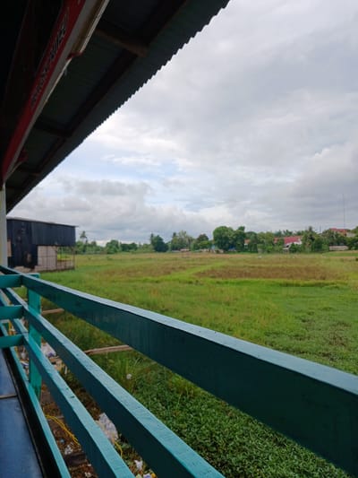 The image depicts an outdoor scene from what appears to be a sheltered vantage point, looking out over a grassy field. In the foreground, a teal-colored wooden railing runs horizontally across the frame, suggesting the perspective is from a balcony, porch, or similar structure. Beyond the railing, a vast expanse of green grass dominates the midground, with patches of dried vegetation interspersed. In the distance, a collection of buildings and trees are visible, hinting at a rural or semi-rural environment. Specifically, a dark-colored, rectangular structure, possibly a barn or warehouse, stands to the left, with a lighter-colored wooden extension. Further back, red-roofed houses and lush green trees are scattered across the landscape under a cloudy sky. The overall atmosphere is overcast and serene. Based on the reference to Geulanggang Teungoh, Indonesia, this scene likely captures a typical view in that region.