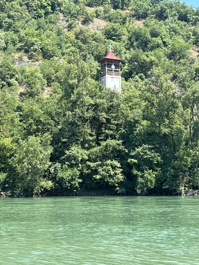 The image depicts a bell tower with a red roof and a cross, nestled amongst lush green trees on a steep hillside. The bell tower is a prominent structure, appearing to be made of stone and wood, with open arches revealing a glimpse of what might be a bell or a statue within. Below the hillside, a calm, greenish body of water stretches across the foreground, reflecting the light and creating a textured surface. The scene suggests a serene natural environment, likely a location overlooking a river or lake, possibly in a rural or semi-rural setting. The specific location in Zvornik, Bosnia and Herzegovina, is not visually identifiable from the image alone, but the presence of a religious structure on a hillside overlooking water is consistent with many scenic locations in the region.