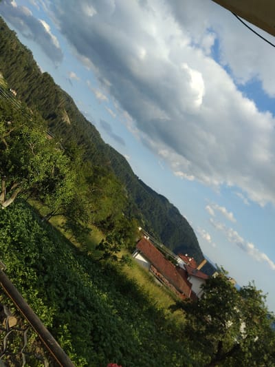 The image depicts a sloped view of a mountainous landscape, likely in or around Momchilovtsi, Bulgaria. In the foreground, there is lush green vegetation, including trees and what appears to be a cultivated field. A dark, weathered metal railing runs diagonally across the lower left of the frame. Further back, nestled on the hillside, are several buildings with orange tiled roofs. The backdrop is a densely forested mountain slope, with the sky above filled with dramatic clouds, ranging from dark, heavy formations to lighter, scattered white clouds against a clear blue sky. The overall impression is of a rural, possibly residential area at the base of a heavily wooded mountain, under a dynamic sky.