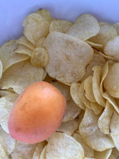 This is a close-up, overhead shot of a pile of potato chips with a single, oblong, orange-pink fruit in the foreground. The fruit is positioned on the left side of the image, partially obscuring the chips beneath it. The chips are golden yellow and appear to be thinly sliced and crispy. The background is a white surface, likely the interior of a container. There are no people or discernible actions taking place. The scene does not provide any specific information that would place it in Osek, Czechia.