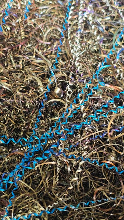 The image displays a close-up, abstract view of metal shavings. The shavings are predominantly metallic bronze and gold in color, with some exhibiting iridescent purple and blue hues, likely due to heat treatment or a coating. Interspersed among these are striking, bright blue, coiled shavings. These blue shavings appear to be the most prominent and visually distinct elements. The texture is complex and chaotic, with countless thin, curled, and twisted pieces of metal creating a dense, layered composition.

The scene is static and appears to be a collection of industrial waste material. There are no people, animals, or natural elements visible. The lighting is somewhat diffused, highlighting the metallic sheen and varied colors of the shavings. The close-up perspective emphasizes the intricate details of the shapes and textures, making it difficult to discern any specific context about where or how these shavings were generated. However, the overall impression is one of industrial byproducts.

There is no discernible text in the image. The location context of Goczałków, Poland, does not provide specific visual cues within the image itself, as it's an abstract representation of material. The image is purely focused on the visual characteristics of the metal shavings.