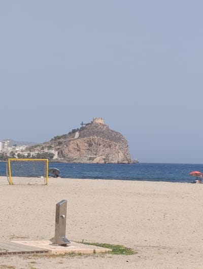The image depicts a wide, sandy beach in Águilas, Spain, with the iconic Castle of San Juan de las Águilas prominently featured on a rocky promontory in the distance. In the foreground, a metallic outdoor shower unit is installed on a wooden platform. Further down the beach, a yellow soccer goal with a net is visible, and beyond that, a blue umbrella shades two people sitting in chairs. The sea is a deep blue, meeting the horizon under a clear, pale blue sky. The background shows a coastal town with buildings nestled against the hillside leading up to the castle. The overall scene suggests a sunny day at the beach, with recreational activities and the historical landmark as key elements of the landscape.