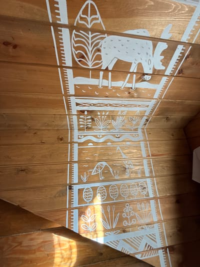 The image shows a wooden ceiling or wall with a white painted mural depicting a stylized natural scene. The mural is divided into horizontal bands, each featuring different elements. The top band shows a large, white moose with antlers, standing amongst abstract trees. Below this, there are depictions of various plants, birds, and what appear to be stylized eggs or pods. The overall style is simple and folk-art-like. The wood is a warm, reddish-brown, with visible grain and knots. Sunlight streams in from the right side of the image, casting shadows and highlighting the texture of the wood. The location is specified as Сосновка, Russia.