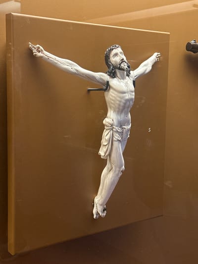 An ivory or similar material sculpture of the crucified Christ (corpus) is displayed within a glass case, resting on a flat, light-brown pedestal. The figure is depicted with outstretched arms and crossed legs, embodying a serene yet pained expression, looking slightly upwards. The sculpture is highly detailed, showing prominent muscle definition, veins, and subtle hints of reddish coloration around the hands, feet, and possibly the forehead, suggesting wounds. The hair and beard are darker, providing contrast to the light body. A dark, metal support structure is visible beneath the torso, holding the figure in position.
The setting appears to be a museum or gallery, with a warm, solid brown background behind the display. Reflections on the glass case are noticeable, including a faint, dark, cylindrical object in the upper left corner of the original image, likely part of the surrounding environment or lighting. A light-colored number "5" is visible on the brown background wall of the display case, possibly an exhibit or inventory number. The overall ambiance is solemn and reverential, fitting for a religious artifact. The location context indicates this piece is in Tepotzotlán, Mexico, suggesting it could be a significant historical or religious artwork from the region or an imported piece housed there.