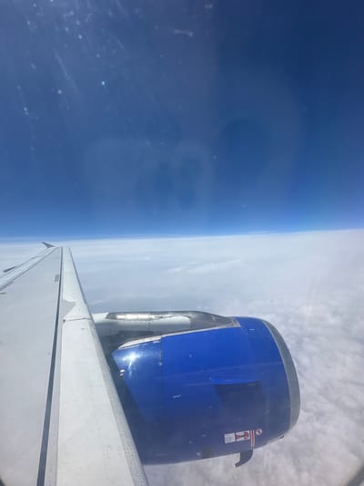An aerial view from an airplane window captures a clear blue sky above a dense layer of white clouds. The wing of the airplane, predominantly white with some metallic detailing, stretches across the left side of the frame. A large, bright blue airplane engine is prominently positioned in the lower right quadrant of the image, with some metallic components visible on its upper surface. The scene is set at an altitude, with no specific location in Viehhausen, Austria, discernible from the image.