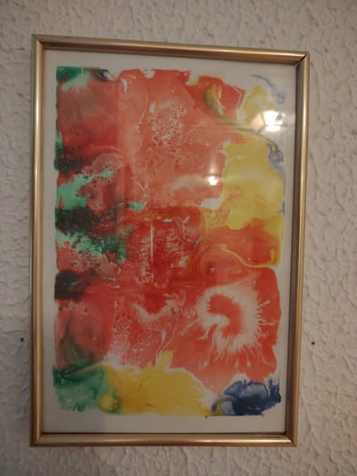 A framed abstract painting with a marbling technique is displayed on a textured white wall. The painting features a vibrant blend of colors, primarily reds, oranges, yellows, greens, and blues, with a swirling, organic pattern. The marbling creates a fluid and dynamic composition, with areas of intense color interspersed with lighter, more diluted sections. Some areas show a frothy, bubbly texture, suggesting the paint may have been manipulated with water or other liquids. The frame is a simple, thin gold-toned metal. The background wall has a rough, stucco-like texture. The lighting appears to be indoor, with a slight glare reflecting off the glass of the frame. The overall impression is one of energetic and abstract expression. There is no visible text in the image. Given the provided location context of Kotka, Finland, this artwork is likely located within a building in that city.