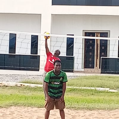 This image captures an outdoor volleyball scene in Koko, Nigeria. In the immediate foreground, a dark-complexioned male, wearing a green and black striped "Dafabet" t-shirt and brown shorts, stands on a sandy court, looking directly forward with a slight smile. Behind a white volleyball net, a second dark-complexioned male, dressed in a red t-shirt and light-colored shorts, is depicted with his right arm extended high, making contact with a yellow and white volleyball above the net. The background features a large, two-story, light-colored building with several dark-tinted rectangular windows and a prominent dark-colored double door. A strip of green grass is visible between the sandy court and a gravel area directly in front of the building. The scene is illuminated by natural daylight.