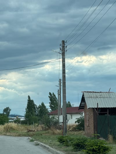The image depicts an outdoor scene in Gelemenovo, Bulgaria, under a cloudy sky. In the foreground, a paved road curves to the left. A small evergreen shrub is planted along the edge of the road. To the right, a rustic brick structure with a corrugated metal roof stands partially visible, featuring a dark green mesh gate. Behind this structure and further back, several trees with green foliage are scattered. Two concrete utility poles with attached wires run vertically through the scene. In the middle ground, a white building with a red roof is visible, along with more trees. In the background, a low structure with a flat roof and some distant buildings or infrastructure are discernible. The sky is filled with heavy, dark clouds, with patches of lighter sky peeking through.