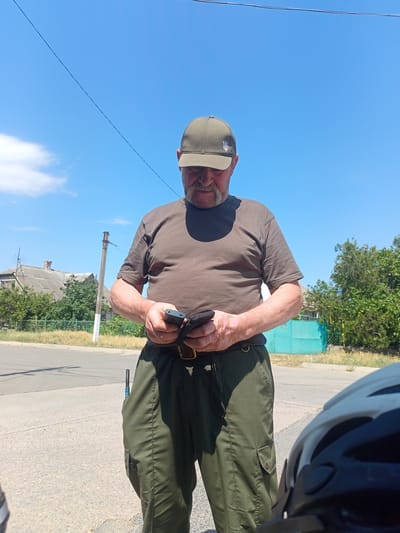 In Shabo, Ukraine, on a bright, sunny day with a clear blue sky and scattered white clouds, a man wearing a dark olive-green baseball cap with a trident emblem and a dark brown t-shirt stands outdoors. He is also wearing dark olive-green cargo pants with a brown belt. The man is looking down at a black mobile phone he is holding in his hands. His right hand is supporting the phone while his left hand is touching the screen. A utility tool with a blue handle is visible sticking out of his left pants pocket. In the background, there is a paved road, a utility pole, some houses with grey roofs, and lush green trees. The lower right corner of the image shows the edge of a white and black helmet, suggesting the presence of a bicycle.